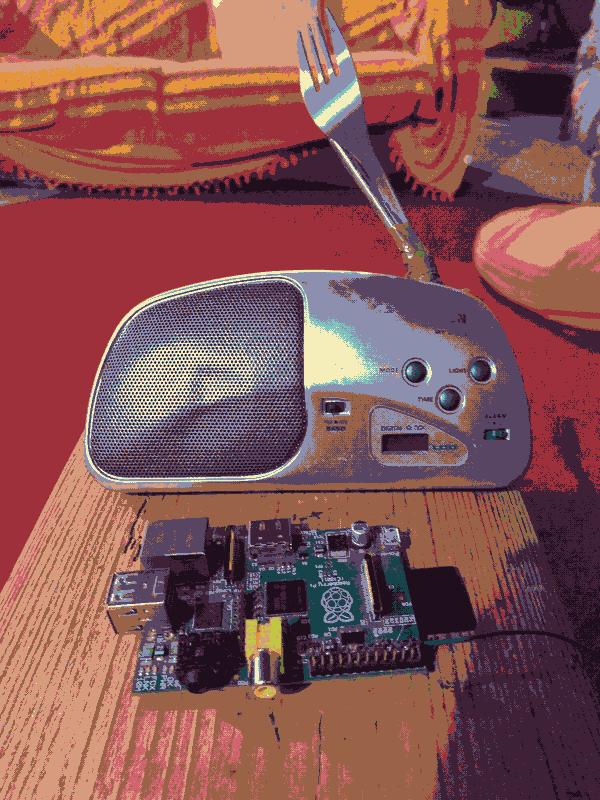 A photo of an old radio, using a fork as an antenna, and a raspberry pi from 2011, on a piece of wood.